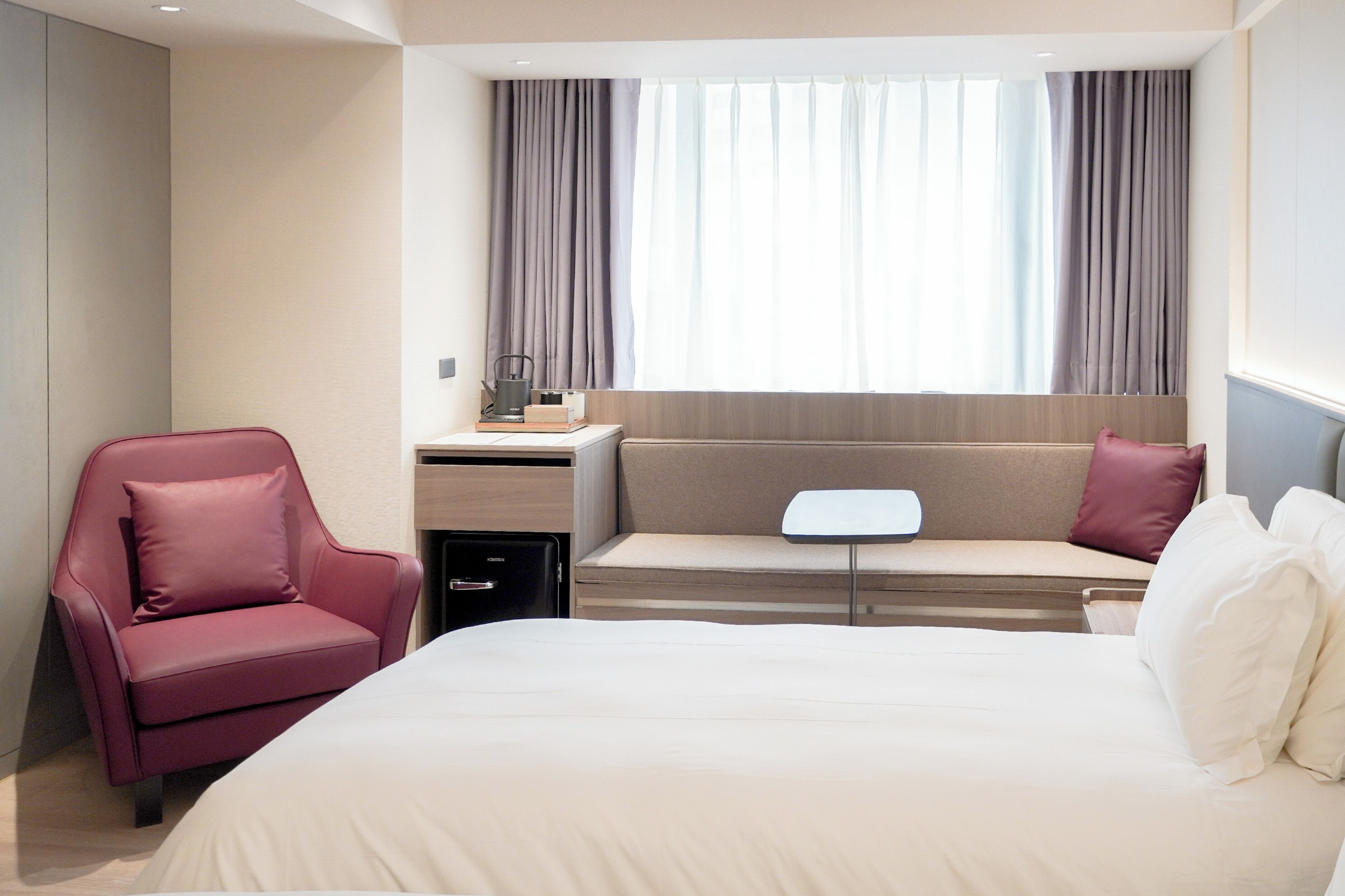 A hotel room with a bed, a red armchair, a small table with a lamp, a sofa, and a window with curtains.