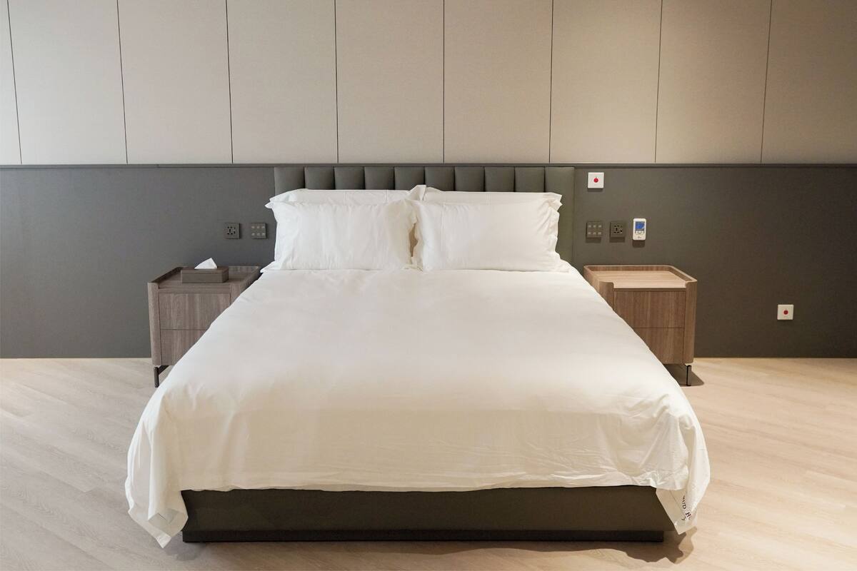 Un lit bien fait, avec une tête de lit grise et du linge de lit blanc, une table de chevet en bois avec une boîte de mouchoirs, et un mur muni d’un interrupteur et d’une prise électrique.