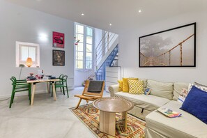 A modern living room with a sofa, a dining table with chairs, a staircase, and a large window.