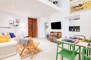 A modern living room with a sofa, a dining table set for two, a bookshelf, and a flat-screen TV mounted on the wall.