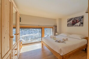 A bedroom with a large window, wooden wardrobe, and a bed with pillows and towels.