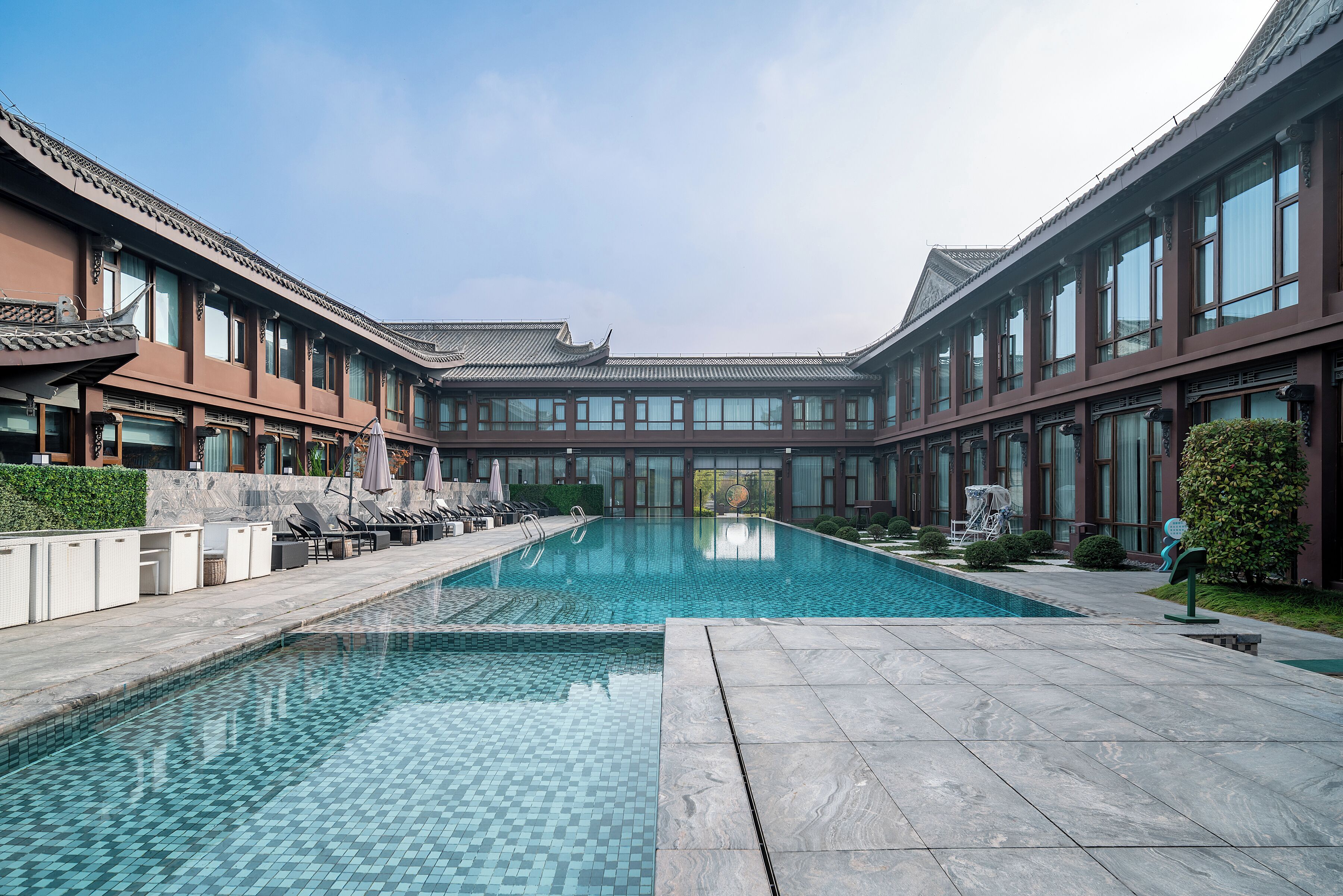 Une piscine dont le sol est recouvert de carrelage, entourée d’un bâtiment doté de grandes fenêtres et d’éléments architecturaux traditionnels.