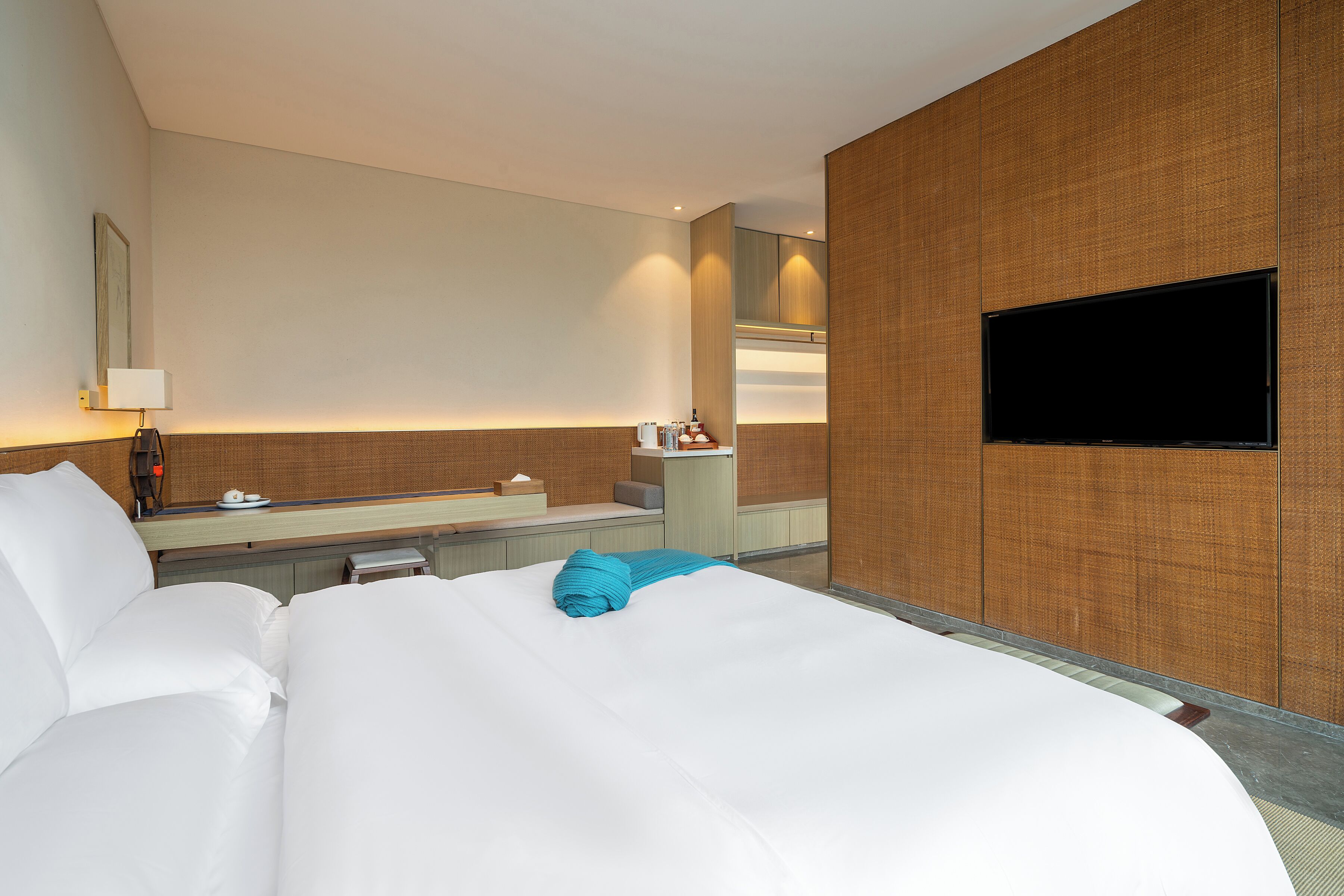 A hotel room with a large bed, a flat-screen TV mounted on the wall, a small desk with a lamp, and a mirror above the desk.