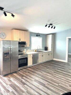 A modern kitchen with stainless steel appliances, light wooden flooring, and a large window.