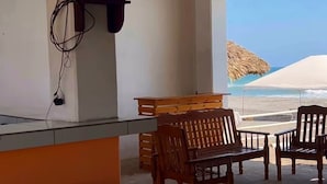 Un bar situé en bord de mer, avec un téléviseur fixé au mur, des chaises en bois et un parasol en chaume.