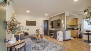 A modern living room with a sofa, coffee table, and decorative plants. A kitchen area with wooden cabinets and a dining table is visible in the background.
