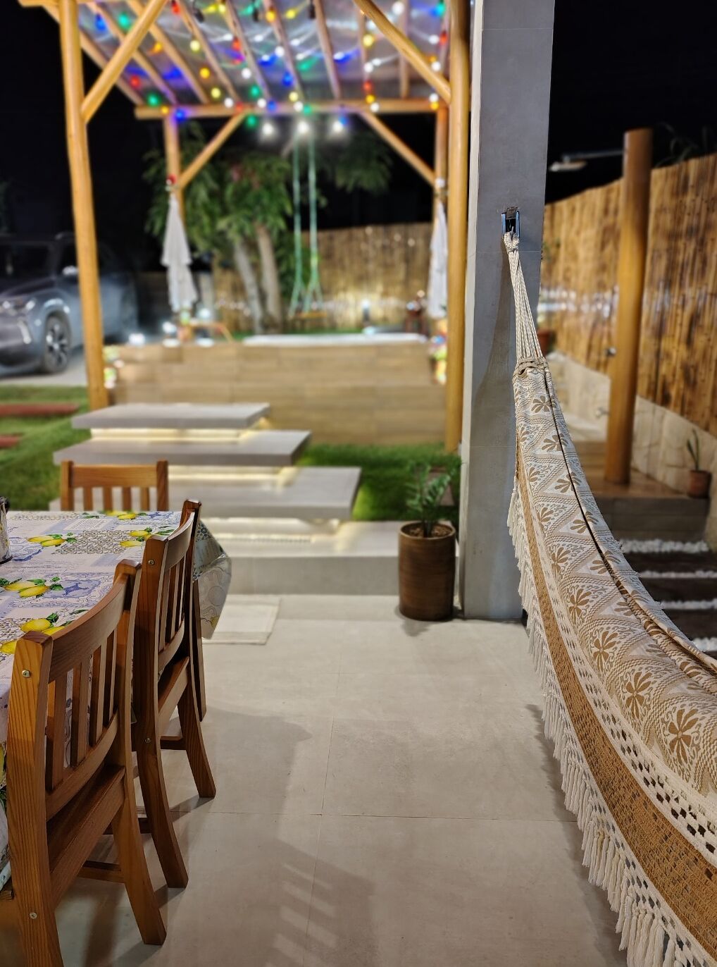 Un espace extérieur aménagé en terrasse, avec des tables et des chaises en bois, une barrière décorative faite de cordes et une pergola illuminée de guirlandes lumineuses.