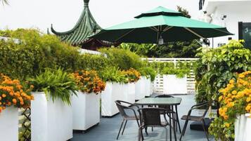 Un patio al aire libre con una mesa y sillas, rodeado de macetas blancas con flores naranjas y una sombrilla verde que da sombra.