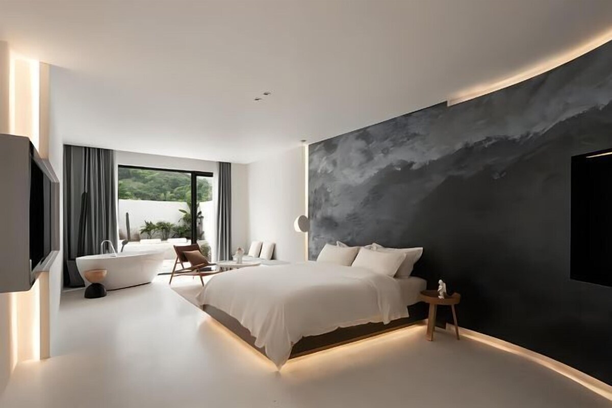 A modern bedroom with a large bed, a wall-mounted TV, and a view of greenery outside.