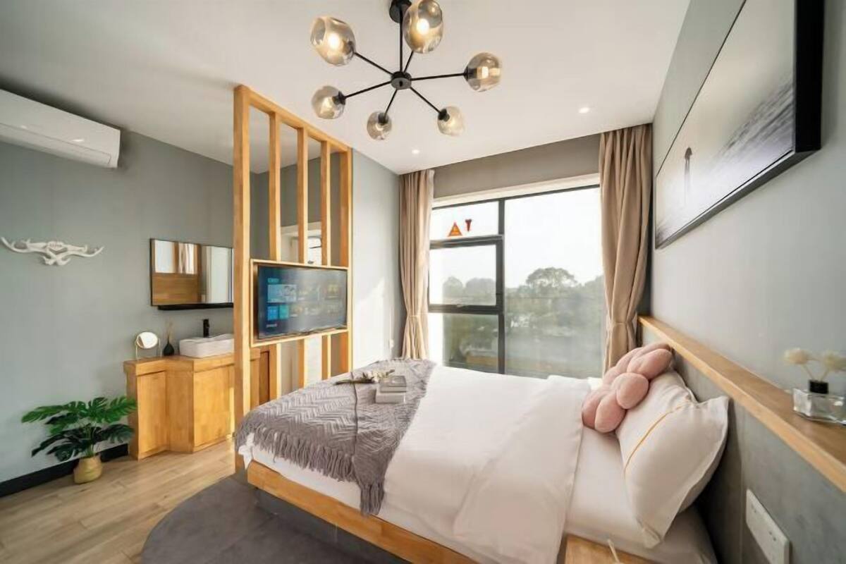 Un dormitorio moderno con cama, televisor, armario de madera, planta y ventana con vistas.