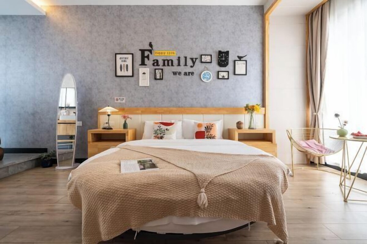A bedroom with a bed, a wall with framed pictures and text, a bedside table with a lamp, and a chair with a vase of flowers.