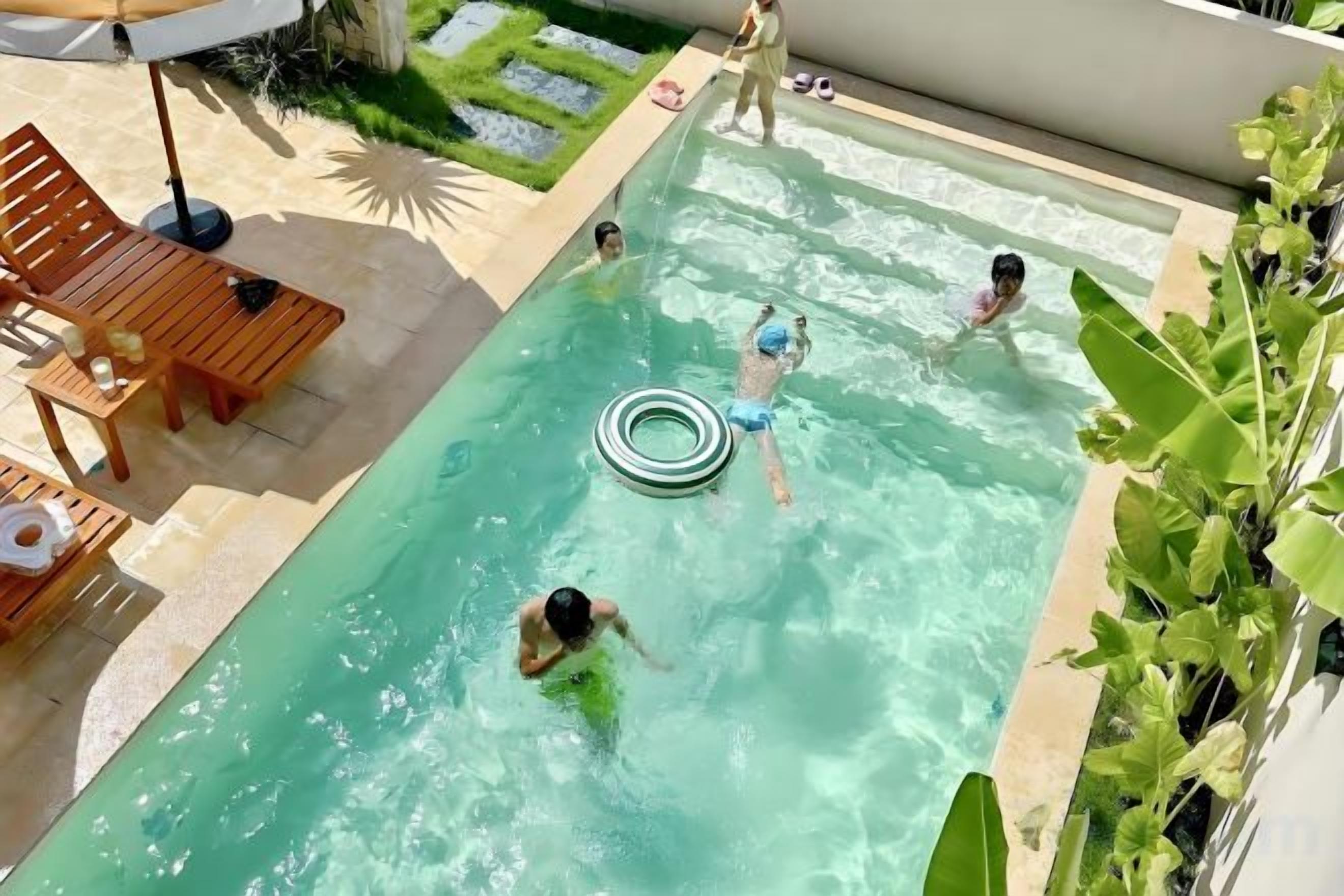 Piscina com pessoas nadando e brincando, rodeada por espreguiçadeiras e um jardim.