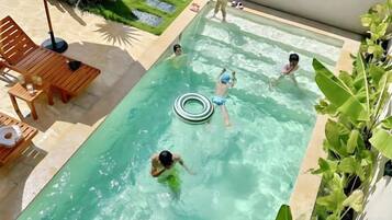 Piscina com pessoas nadando e brincando, rodeada por espreguiçadeiras e um jardim.