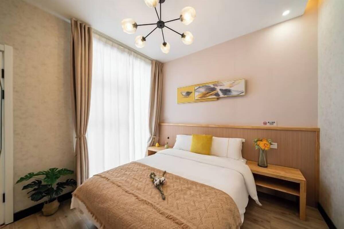 Habitación de hotel con cama, mesita de noche, lámpara, cortinas, un cuadro en la pared y una planta en maceta.