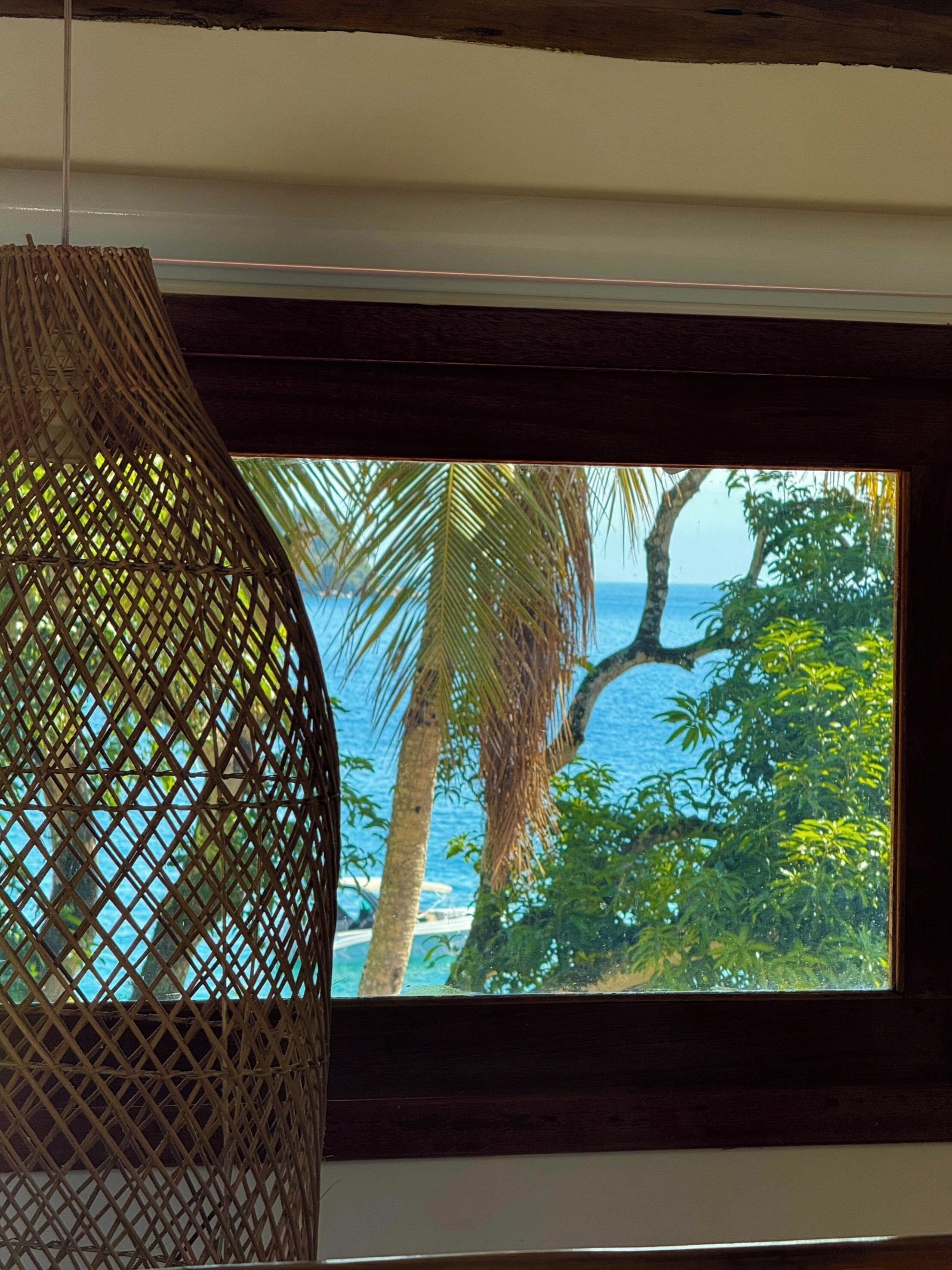 A room with a view of the ocean through a window, a woven hanging light fixture, and a wooden ceiling.