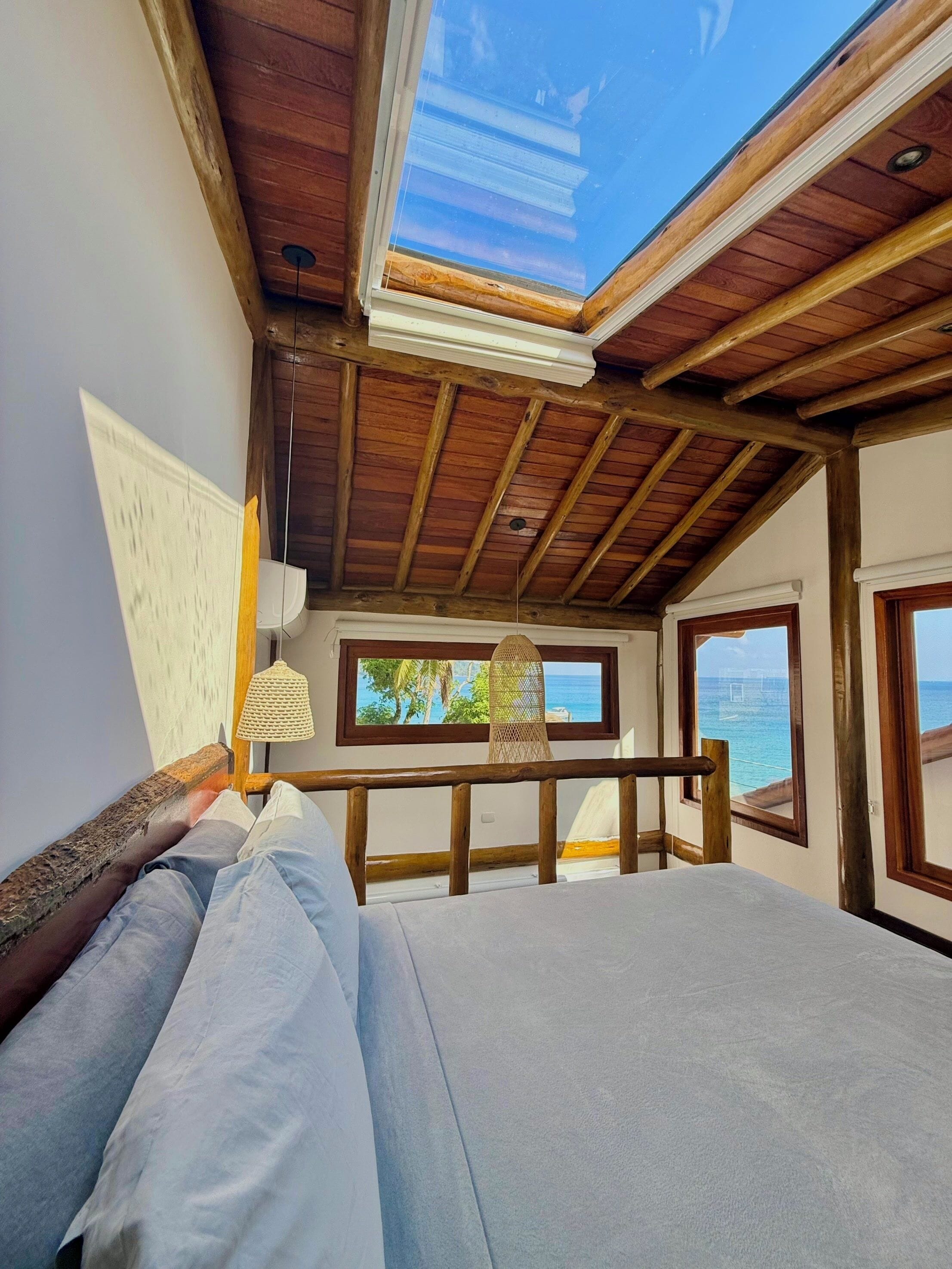 A bedroom with a wooden ceiling, a bed, a window with a view of the sea, and a hanging light fixture.