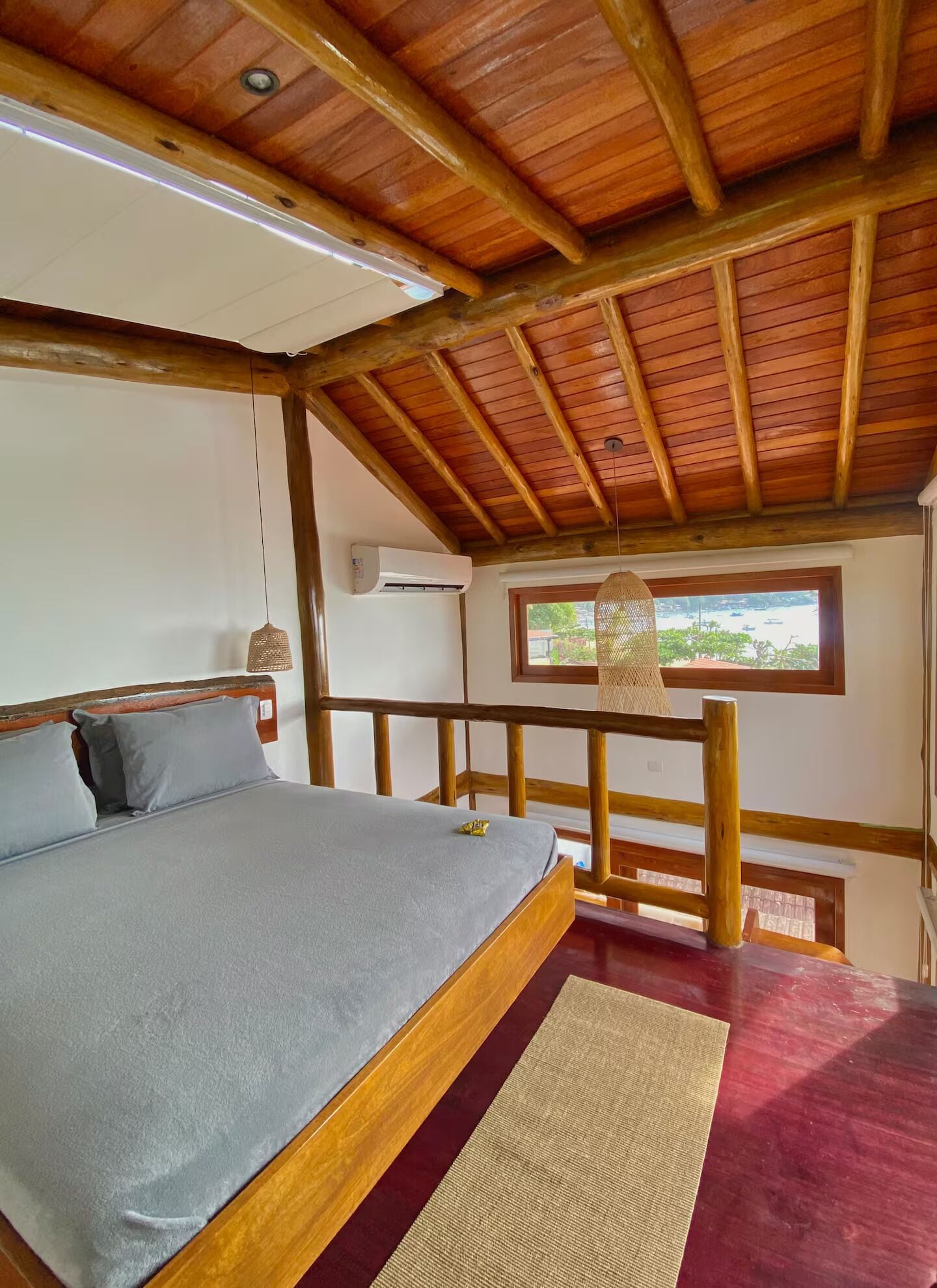 A bedroom with a wooden ceiling, a bed with grey bedding, a window with a view, and a balcony railing.