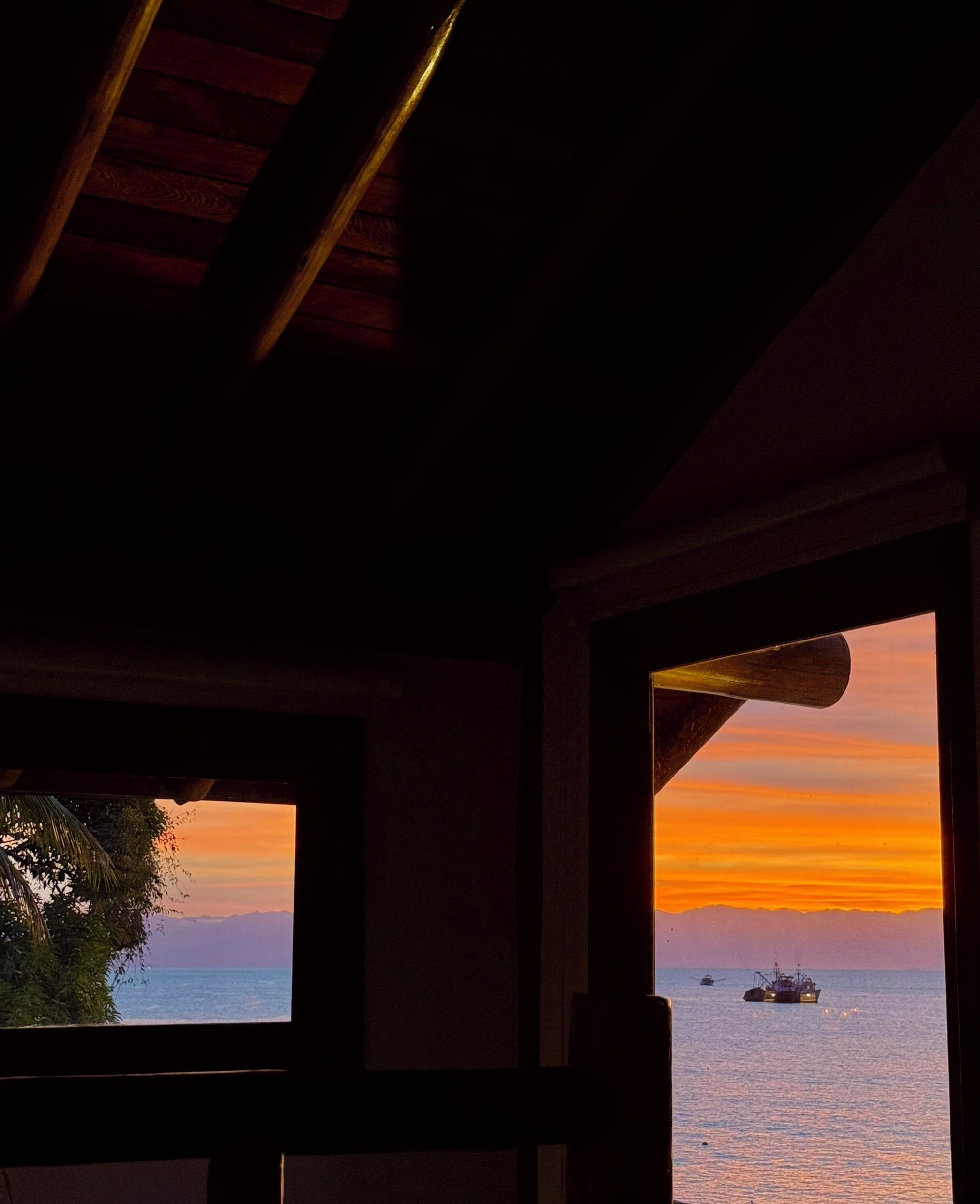 Vista desde el interior de un edificio con techo de madera y dos ventanas. Una ventana muestra una puesta de sol sobre el océano con un barco a lo lejos.