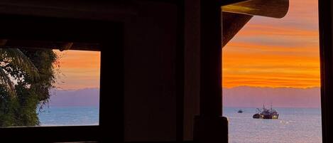 Vista desde el interior de un edificio con techo de madera y dos ventanas. Una ventana muestra una puesta de sol sobre el océano con un barco a lo lejos.