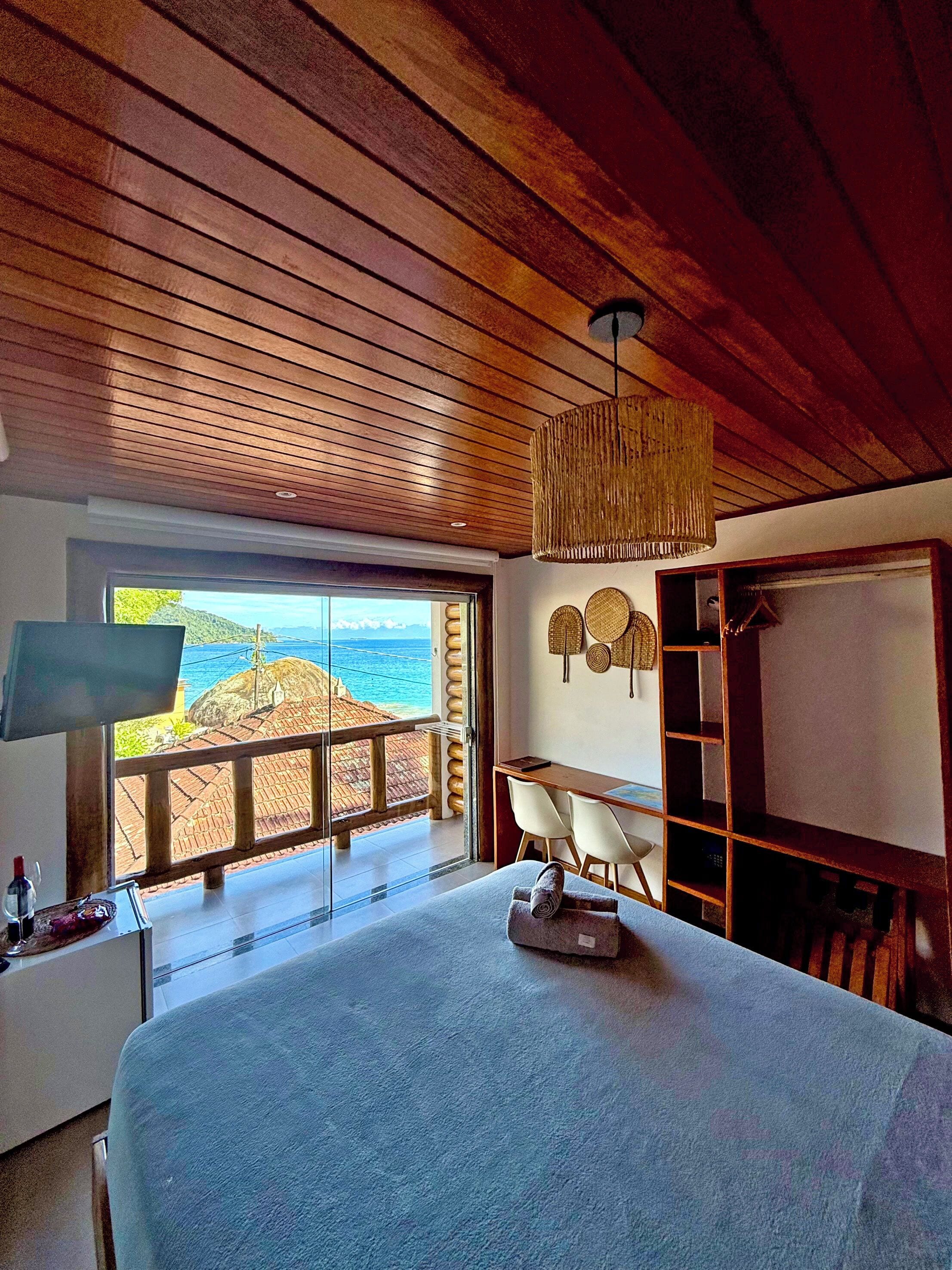 A room with a large bed, a desk, and a TV. There is a view of the ocean through a large window.