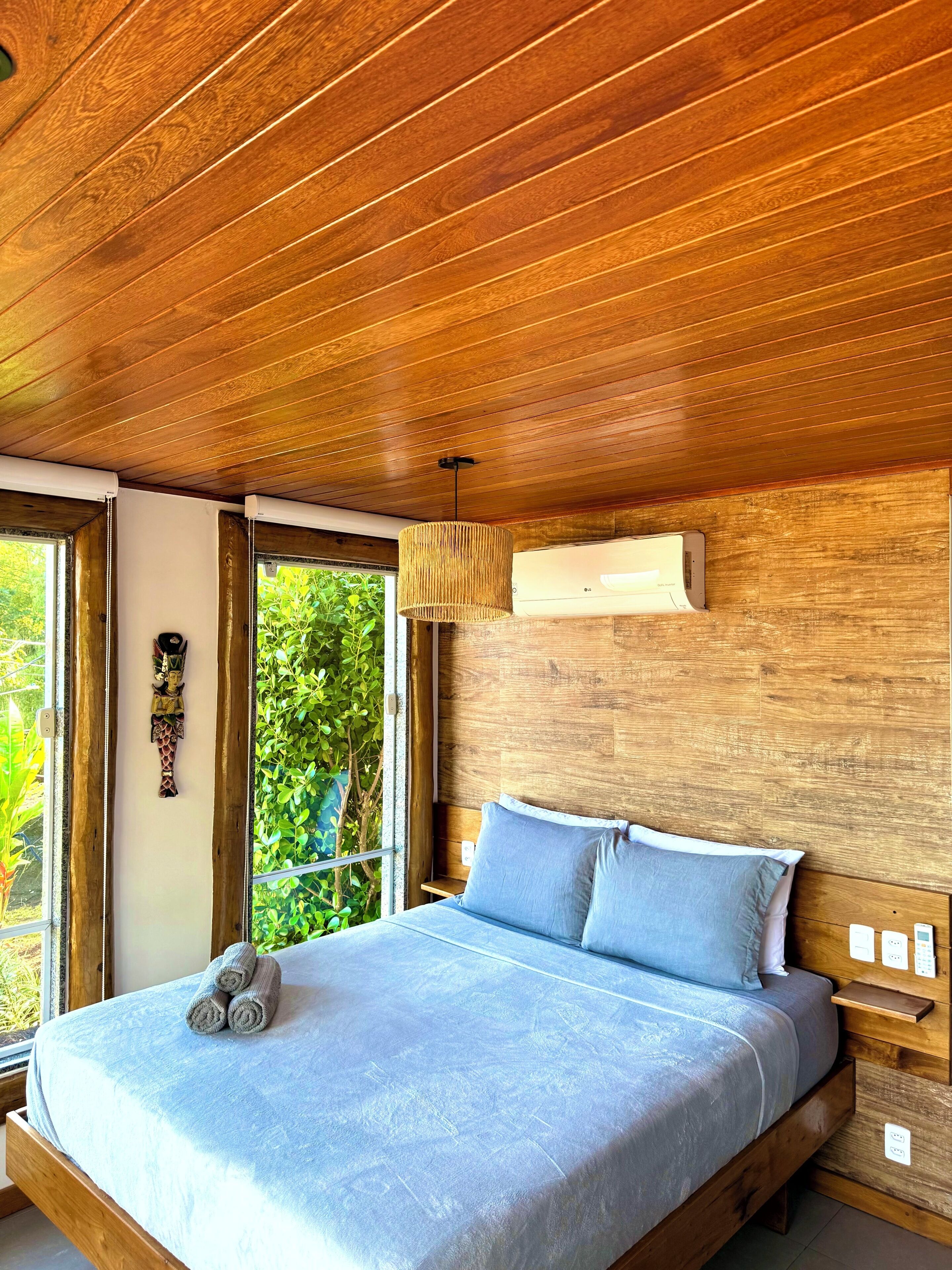 A bedroom with a wooden ceiling, a bed with blue bedding, a hanging light fixture, and a window with a view of greenery.