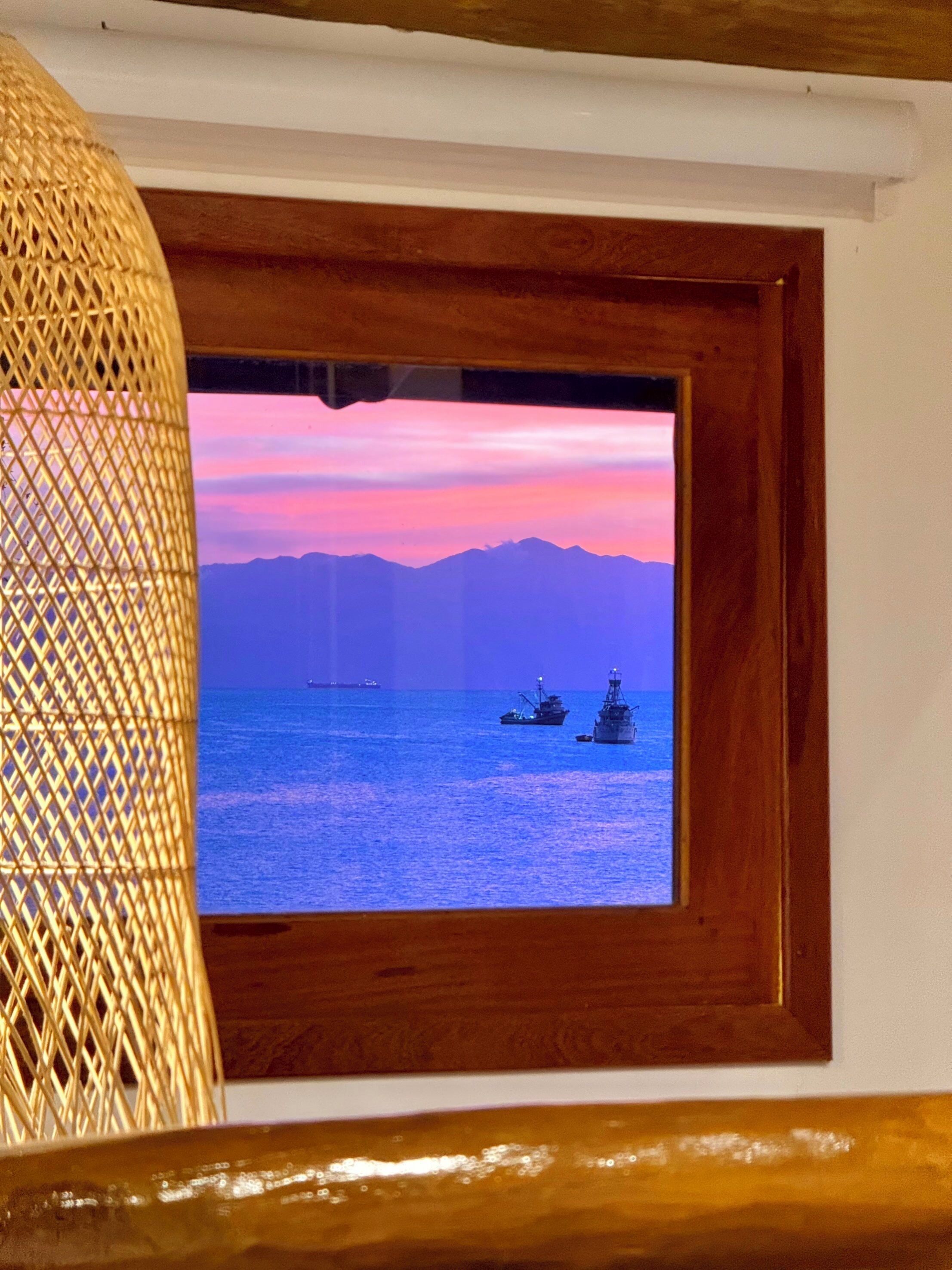 Una vista enmarco de un atardecer sobre el océano con barcos, enmarcada por un borde de madera y un objeto de mimbre.