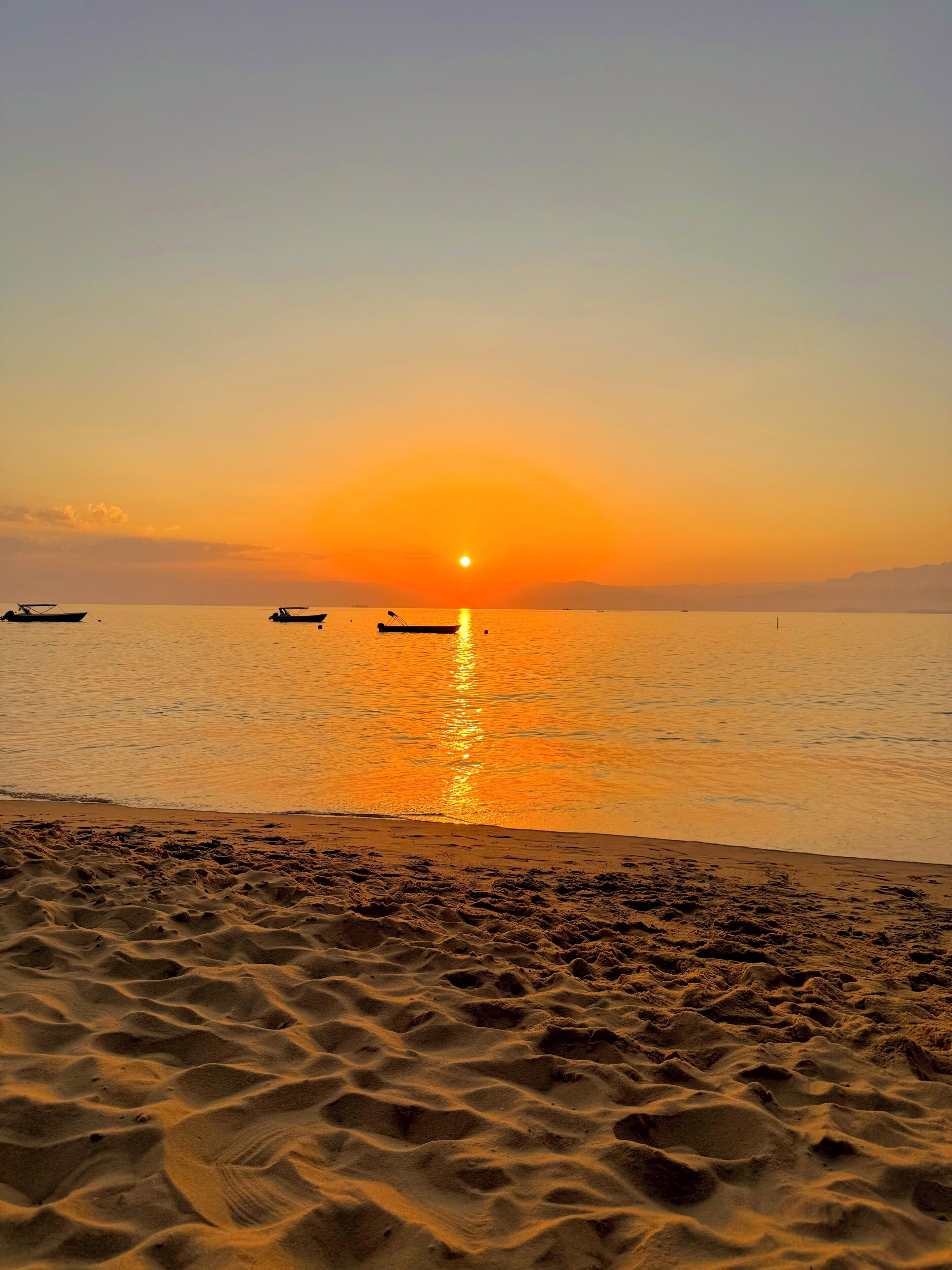 A beach at sunset with boats on the water and sand in the foreground.