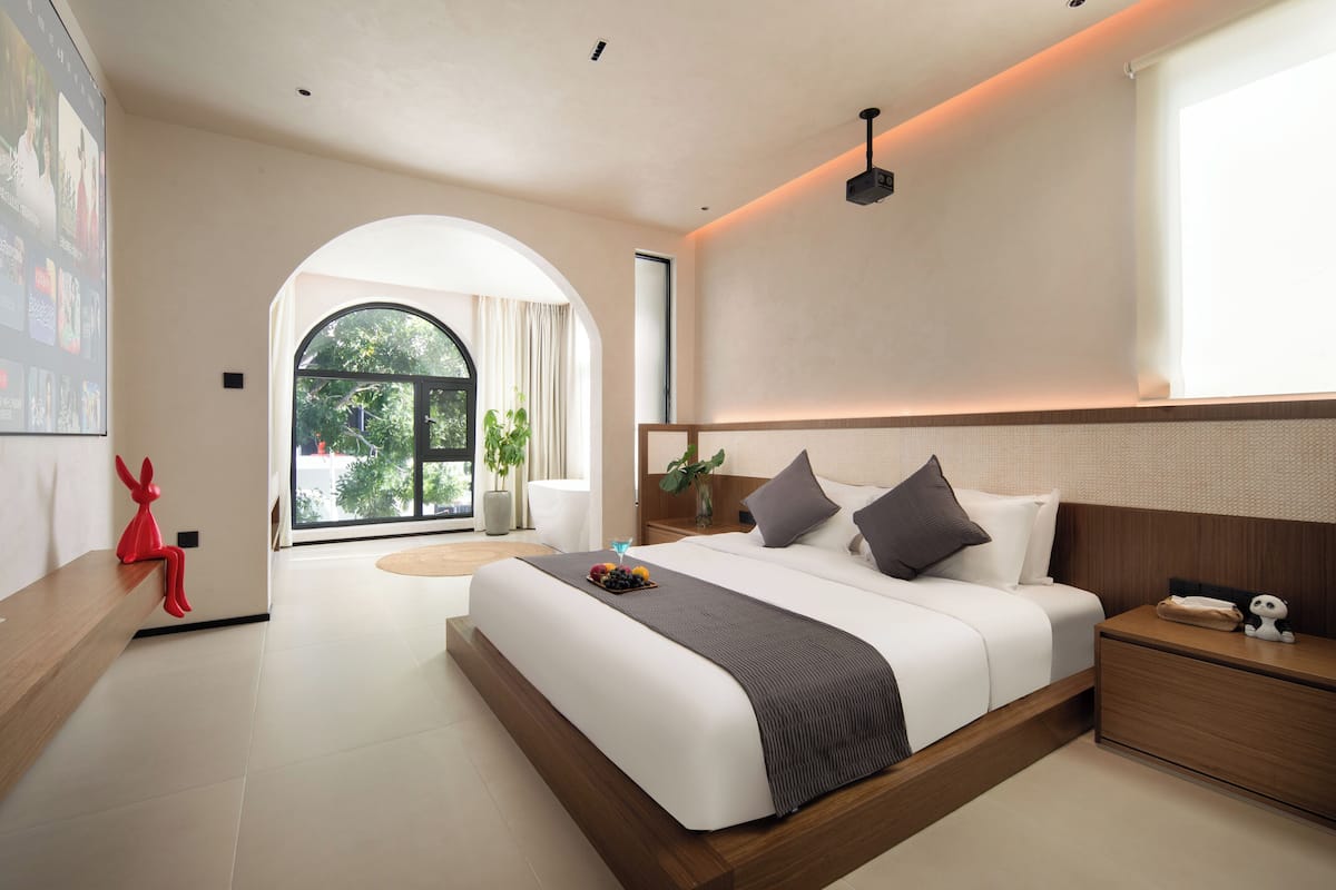 Una habitación de hotel moderna con una cama grande, una mesita de noche, un objeto decorativo rojo y vistas al exterior a través de un ventanal.