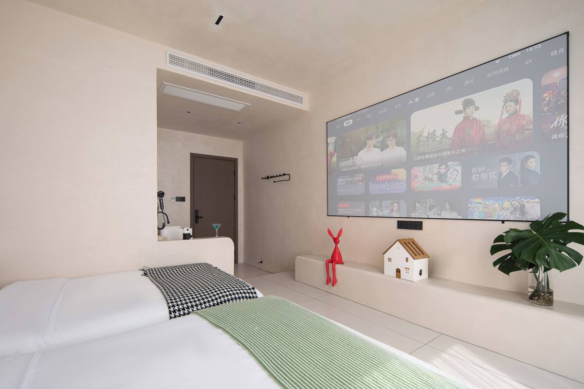 Una habitación de hotel moderna con un televisor de gran tamaño montado en la pared mostrando la interfaz de un servicio de streaming, una cama con ropa de cama estampada, una pequeña decoración con forma de casa y una planta en maceta.