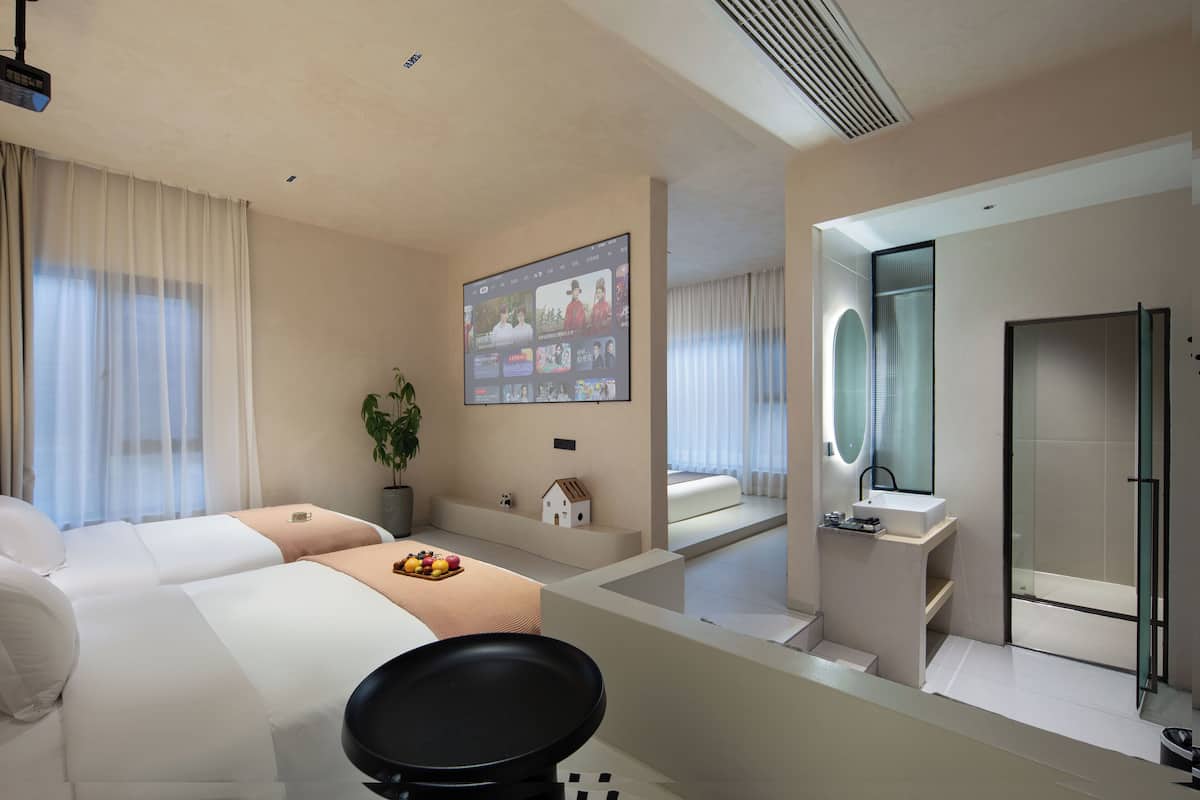 Una habitación de hotel moderna con una cama grande, un sofá pequeño, una mesa de comedor con fruta, una pantalla de televisión, un baño con ducha de vidrio y un espejo grande.