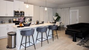 Et moderne køkken med hvide skabe, en central ø med barstole, en sort sofa og et spiseområde med et hvidt bord og stole.