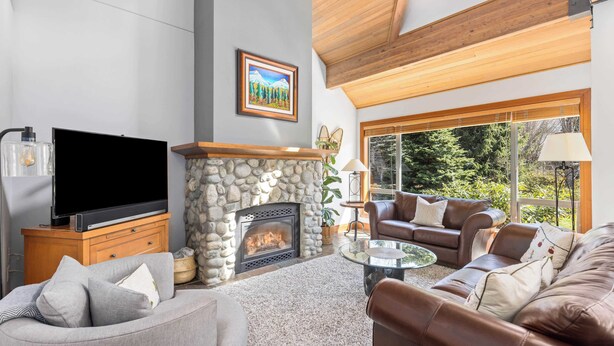 A living room with a stone fireplace, leather sofas, a flat-screen TV, and a large window with a view of trees.