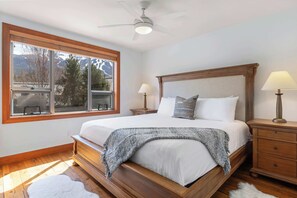 Une chambre à coucher avec un grand lit, une tête de lit en bois, deux tables de chevet avec des lampes, une fenêtre donnant sur des arbres et des montagnes, et un ventilateur de plafond.