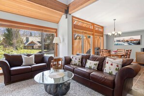 A spacious living area with a brown leather sofa, a glass coffee table, and a dining area with wooden furniture. - Village North townhouse w/ hot tub (Whistler)