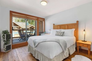 A bedroom with a large bed, a nightstand, a lamp, a sliding glass door leading to an outdoor area with a chair and table. - Village North townhouse w/ hot tub (Whistler)