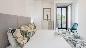 A modern bedroom with a large bed, a grey headboard, a bedside table with a mirror, a blue chair, and a view of the outside through a glass door. - GuestReady - Rose Apartment (Porto)