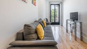 A modern living room with a long grey sofa, a glass coffee table, and a television on a stand. - GuestReady - Rose Apartment (Porto)