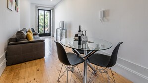 A modern living room with a glass dining table, black chairs, a grey sofa with cushions, a TV on a stand, and a glass door leading outside. - GuestReady - Rose Apartment (Porto)