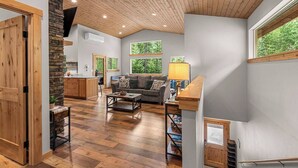 A spacious living area with a wooden ceiling, large windows, a sofa, a coffee table, and a kitchen area visible through an open door. - Eagle Creek Hideaway (Leavenworth)
