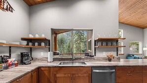 A modern kitchen with wooden cabinets, a marble countertop, and a large window offering a view of trees. - Eagle Creek Hideaway (Leavenworth)