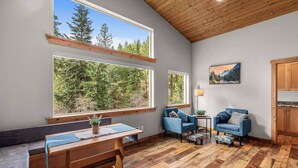 A cozy room with a wooden ceiling, large windows with forest views, a bench, two blue armchairs, a small table, and a painting on the wall. - Eagle Creek Hideaway (Leavenworth)