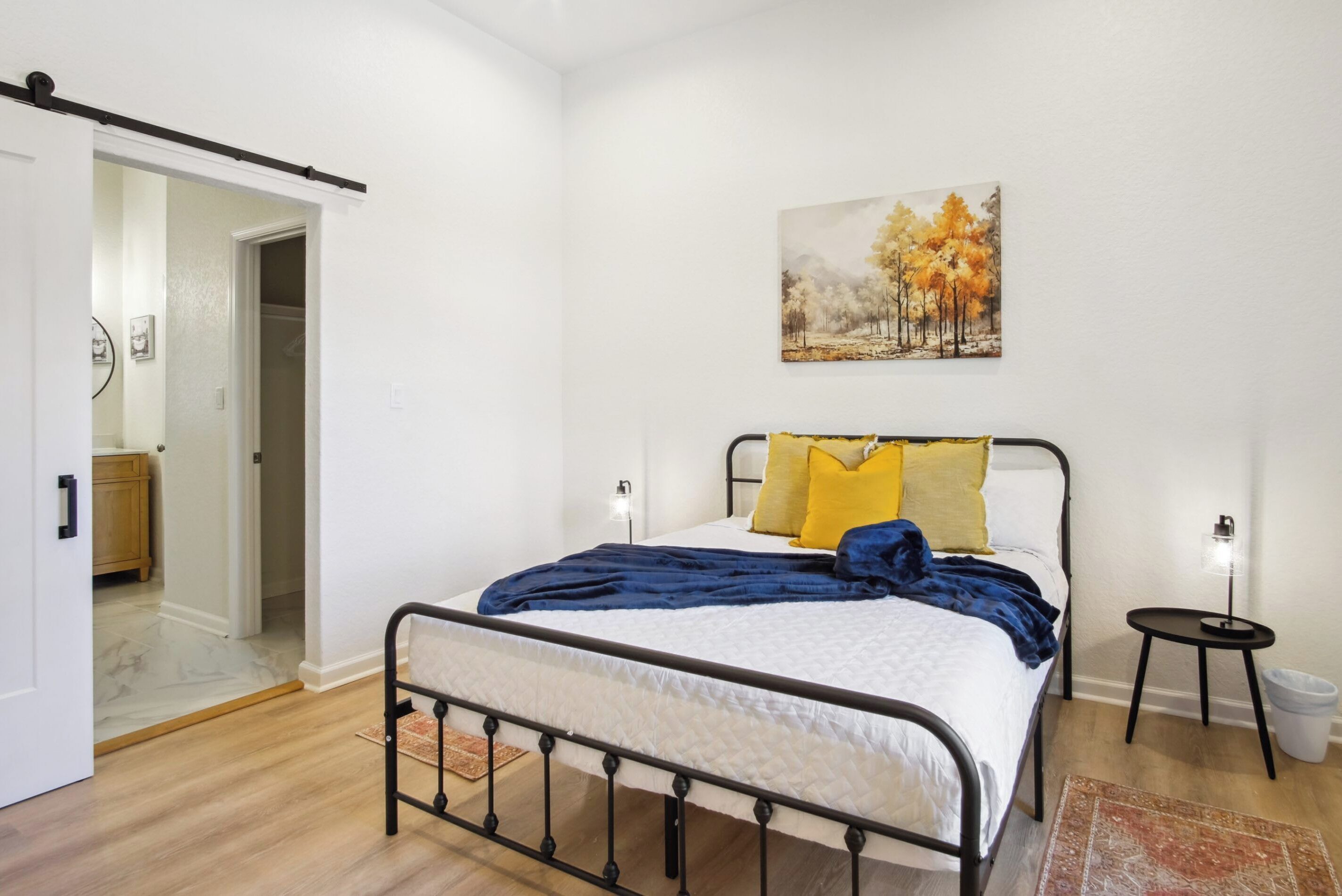 A bedroom with a bed, a painting, a bedside table, and a door leading to another room.