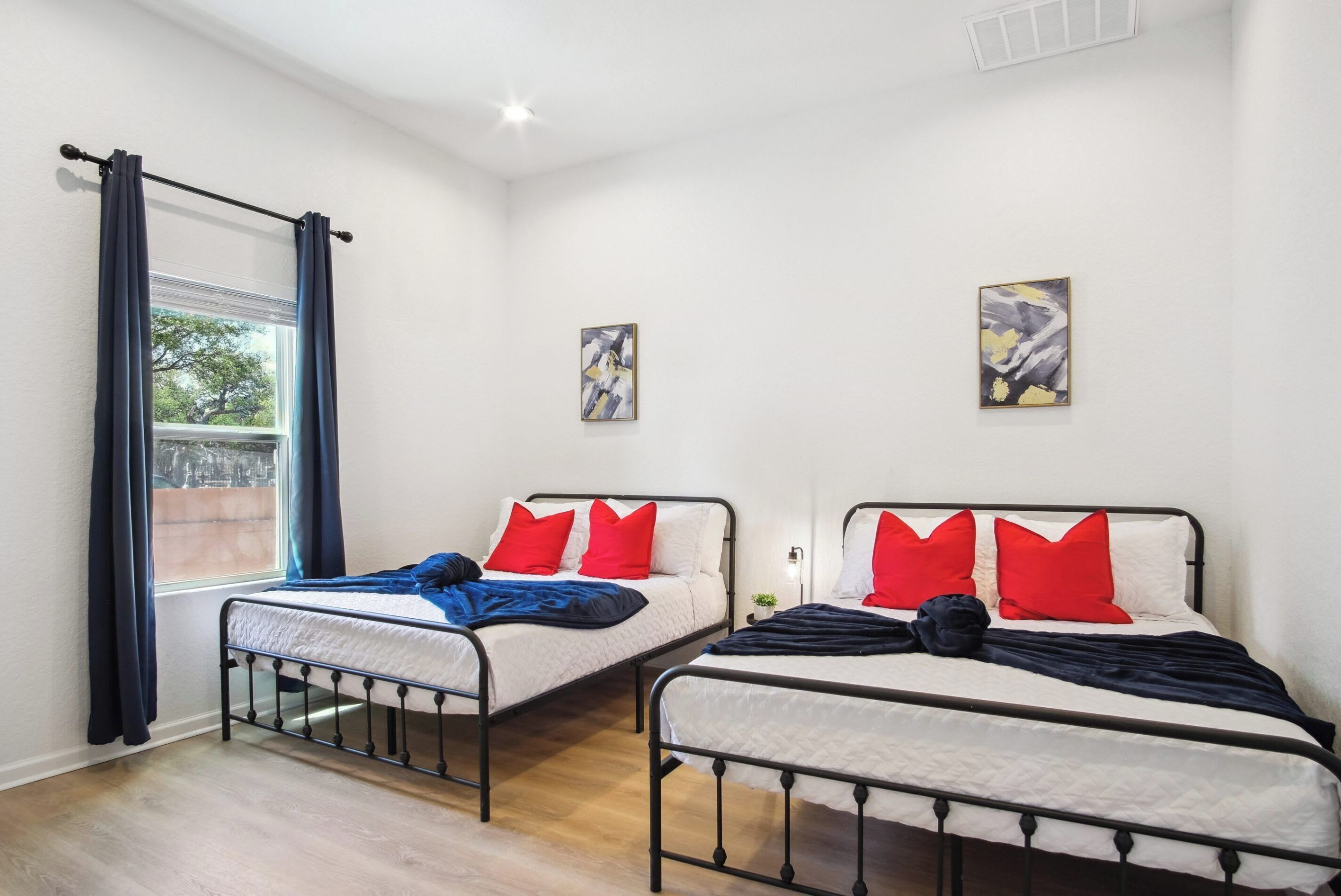 A room with two metal beds, white bedding, red pillows, and blue curtains. There are two paintings on the walls.