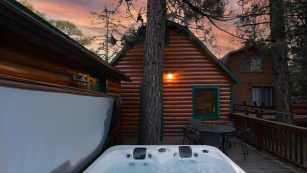 A log cabin with a hot tub on a deck, surrounded by trees and a sunset.