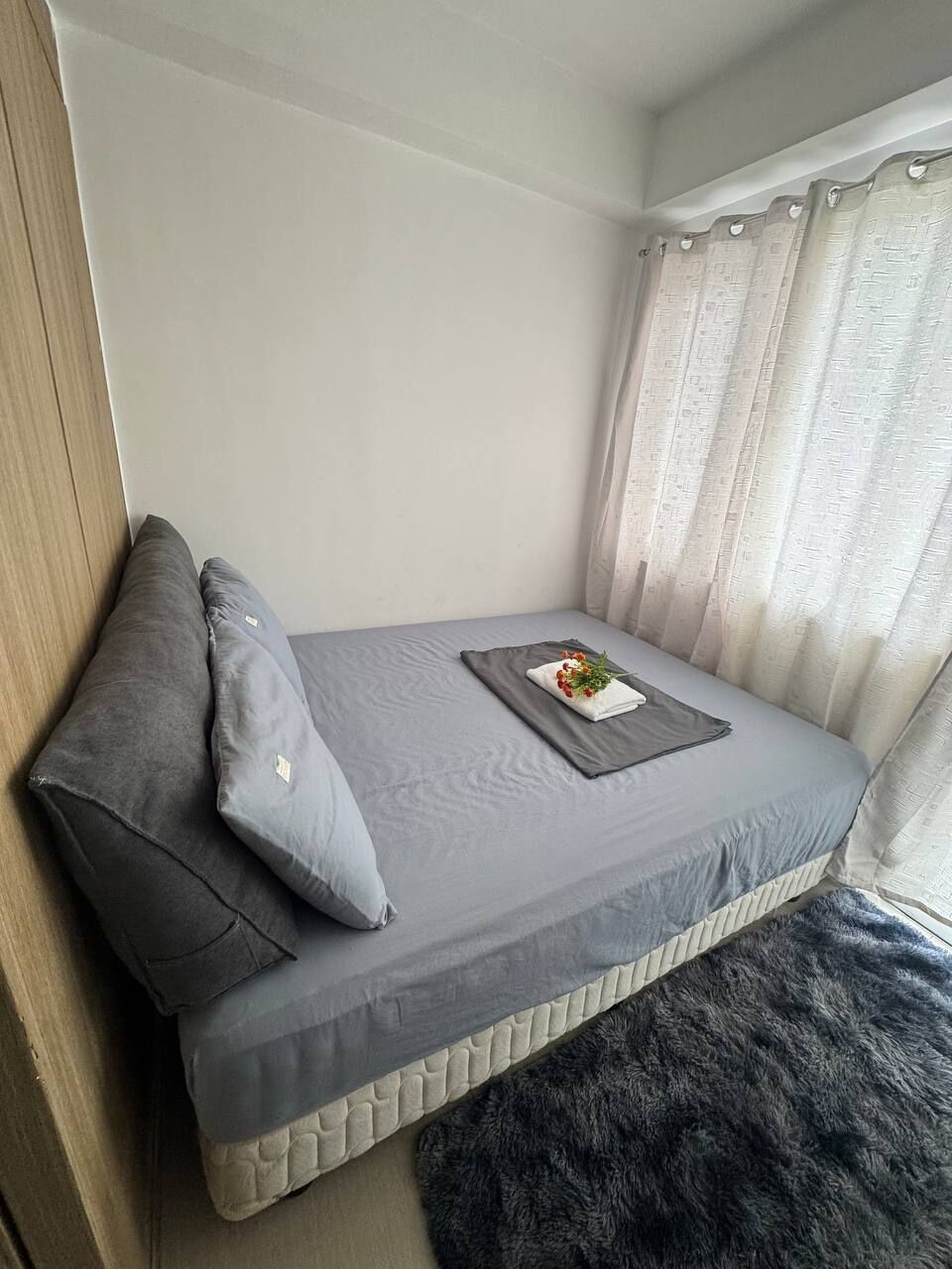 Uma cama pequena e bem arrumada com roupa de cama e almofadas cinzas, um tapete felpudo escuro e uma janela com cortinas finas.
