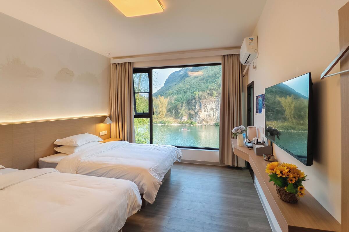 A hotel room with two beds, a large window with a view of a lake and mountains, a flat-screen TV, and a wooden bedside table with a vase of flowers.