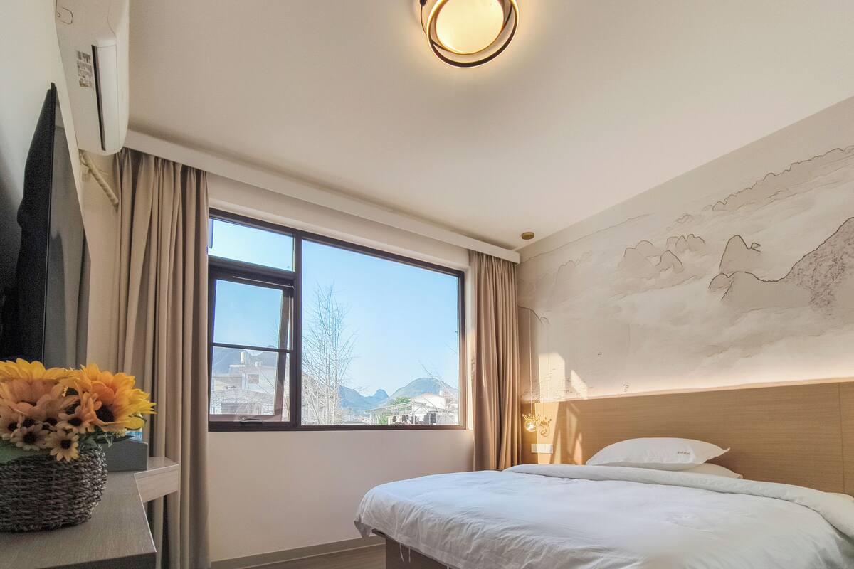 A hotel room with a bed, a window with curtains, a wall-mounted air conditioner, and a decorative wall piece.