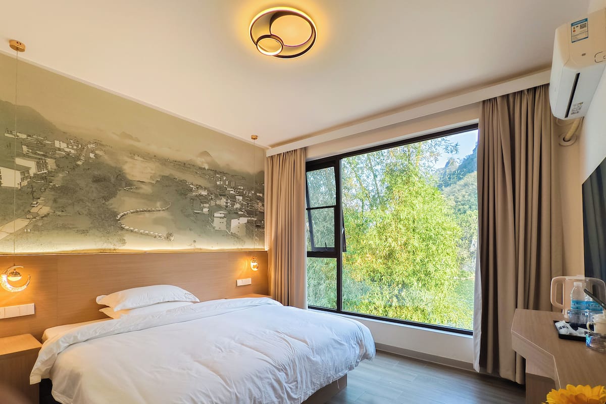 A hotel room with a large bed, a mural on the wall, a window with a view of greenery, and a desk with a water bottle and a cup.