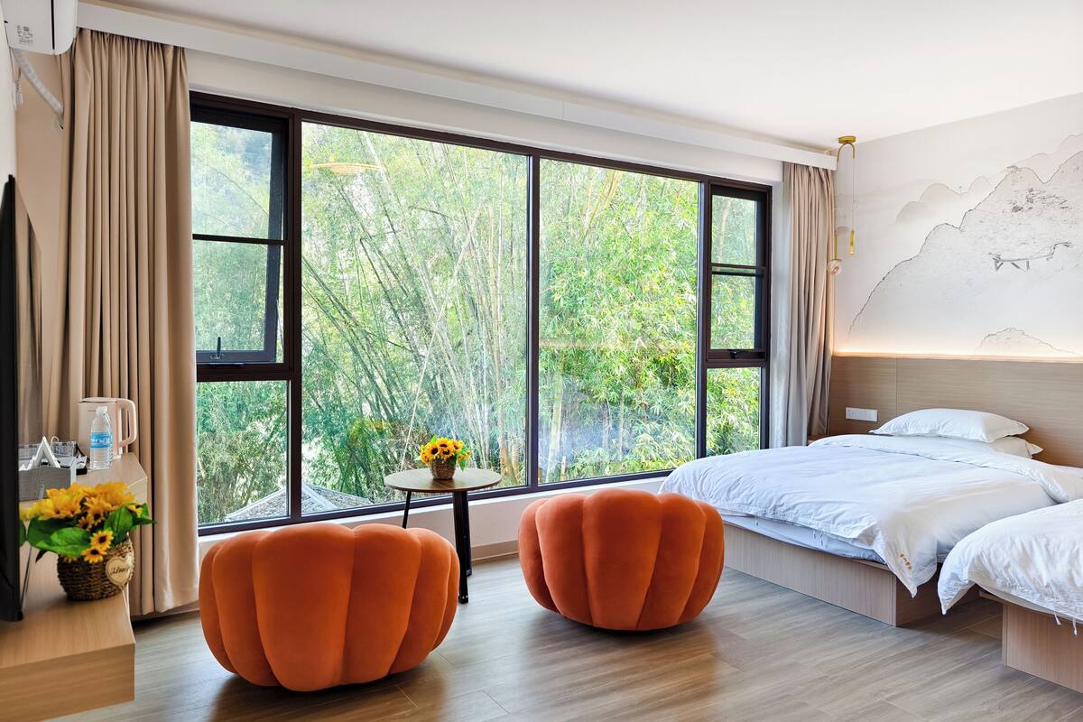 A modern hotel room with a large window, a bed, two orange armchairs, a small table with a vase of flowers, and a water bottle on the nightstand.