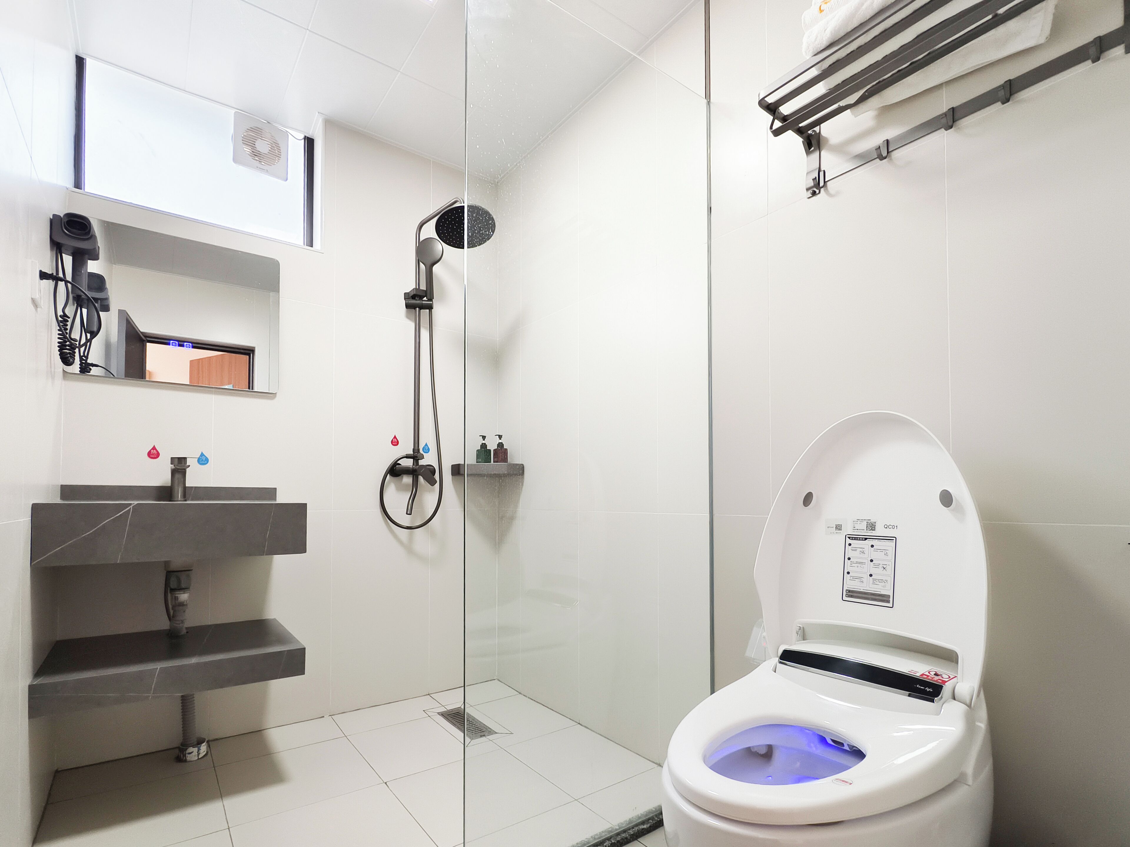 A modern bathroom with a glass-enclosed shower, a white toilet with a blue light, and a grey sink with a towel rack above it.